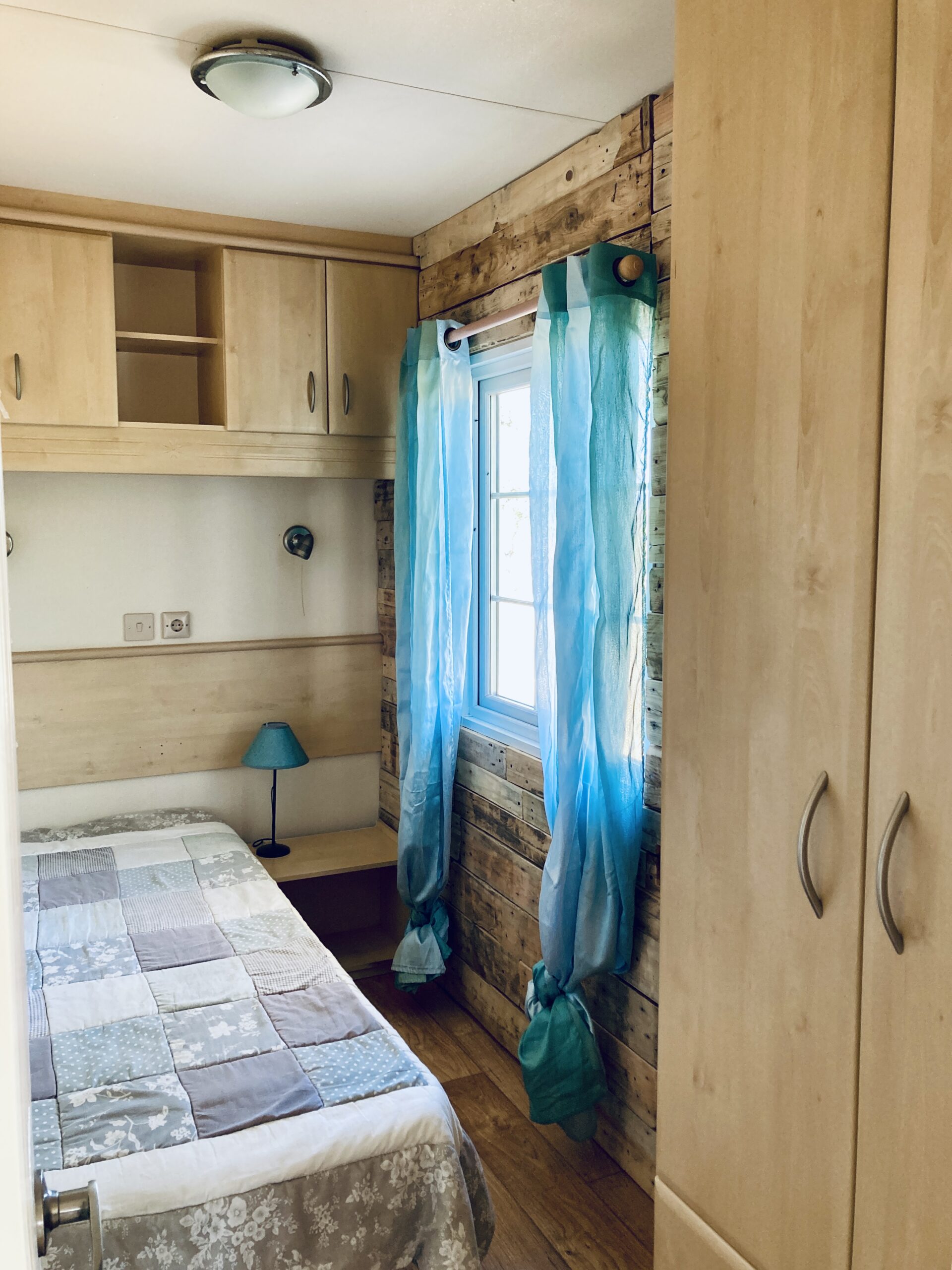 Look into the single bedroom with light blue curtains of a mobile holiday cottage at Equus Ourique