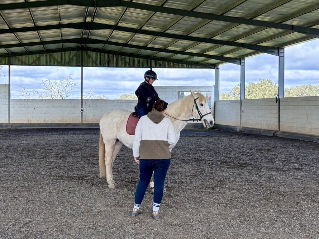 Woman on a white horse speaking to her trainer in an indoor school