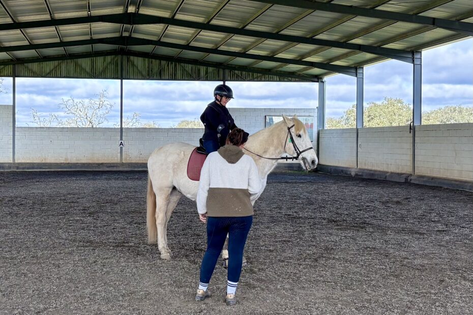 Woman on a white horse speaking to her trainer in an indoor school